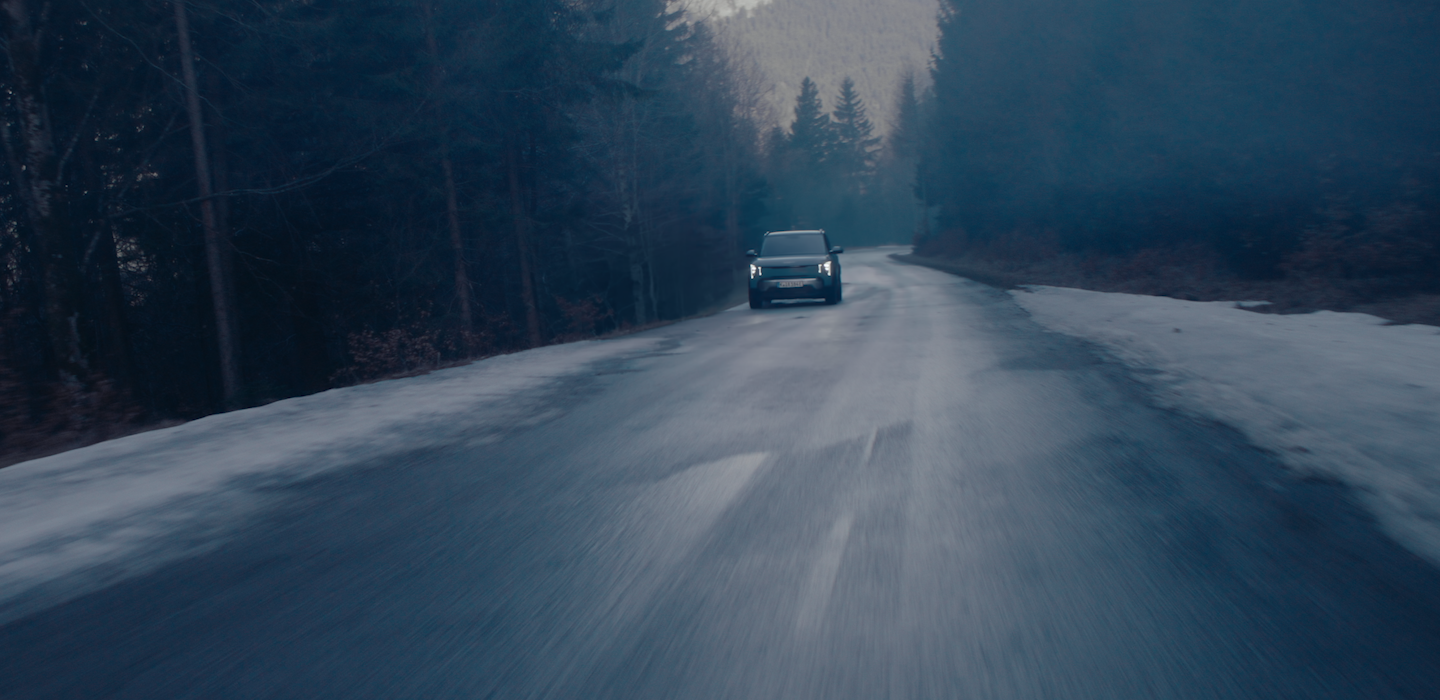 Dunkle, atmosphärische Aufnahme eines Kia Fahrzeugs, das bei Dämmerung mit eingeschalteten Scheinwerfern auf einer nassen Waldstraße auf die Kamera zufährt. Entlang der Straße liegen vereinzelt Schneereste.